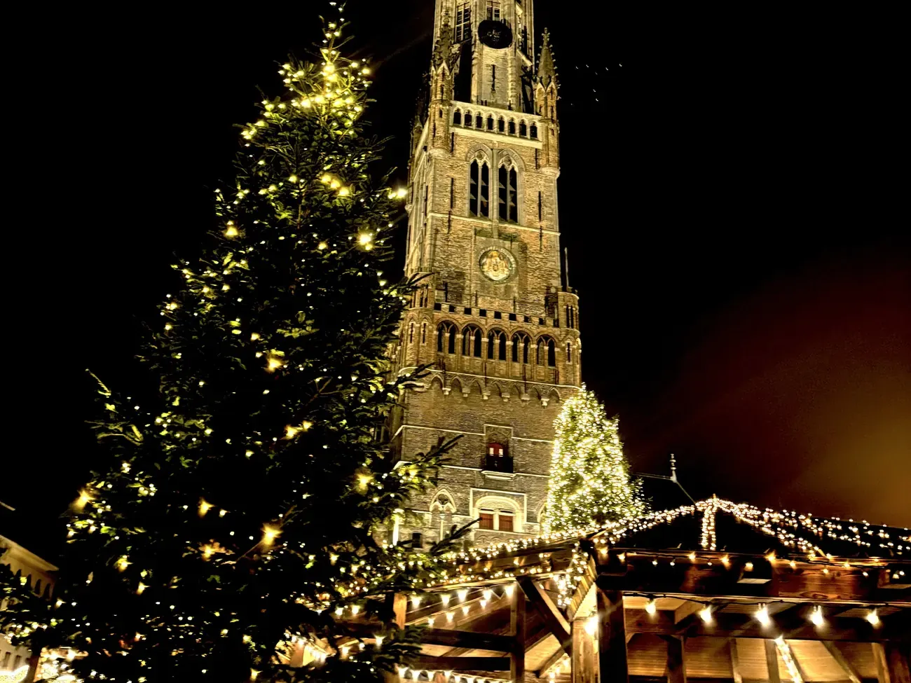 The Belfry of Bruges lit at night above a Christmas tree and a market chalet strung with lights on the Markt