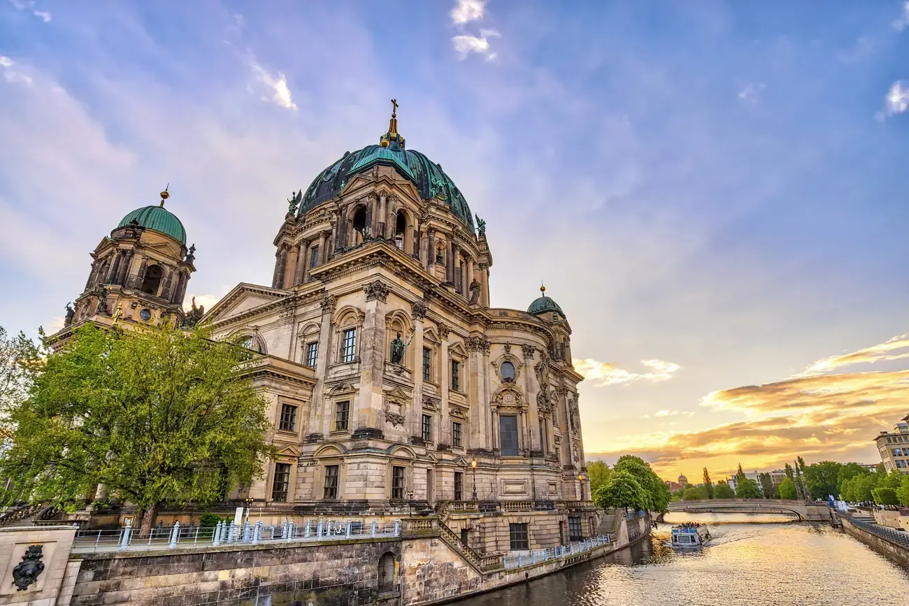 Berlin Cathedral rising above the Spree riverbank at sunset, with a boat passing below