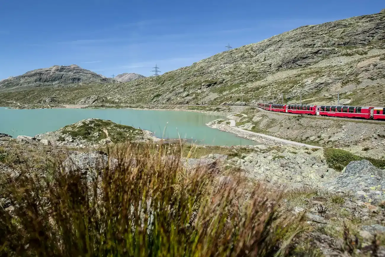 Distant Bernina Express running beside a turquoise lake on the rocky Bernina Pass