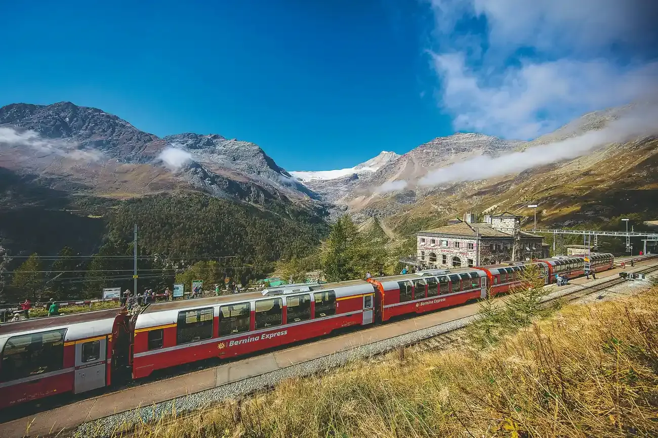 Bernina Express passing a stone mountain station with clouds over the Alpine valley