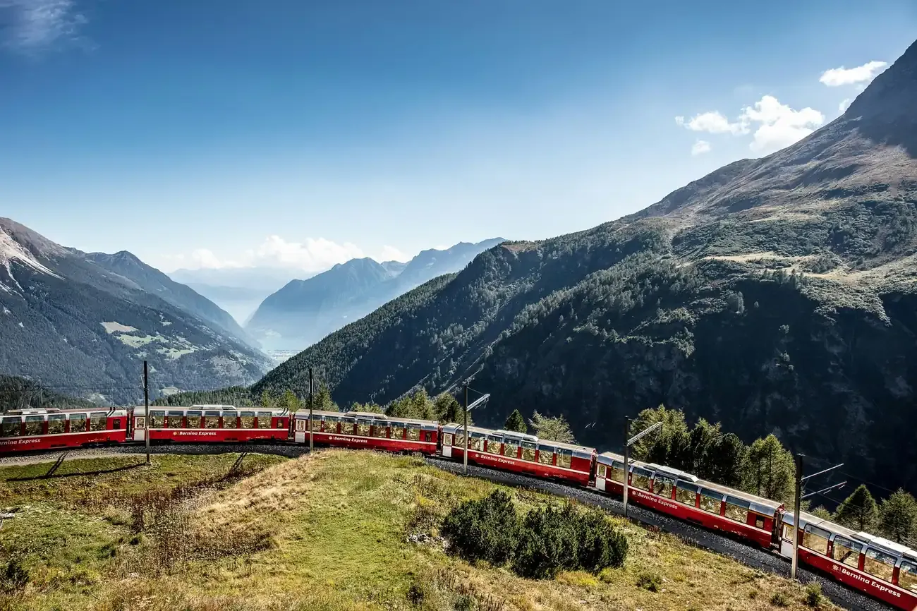 Long red Bernina Express winding along a green Alpine hillside with mountains beyond