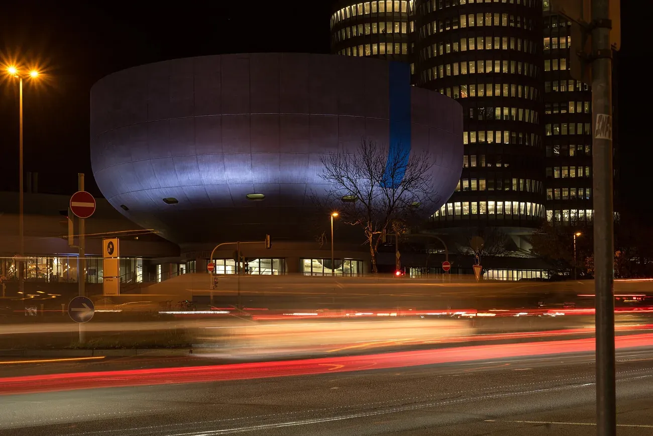 The bowl-shaped BMW Museum lit at night with the four-cylinder BMW tower behind in Munich