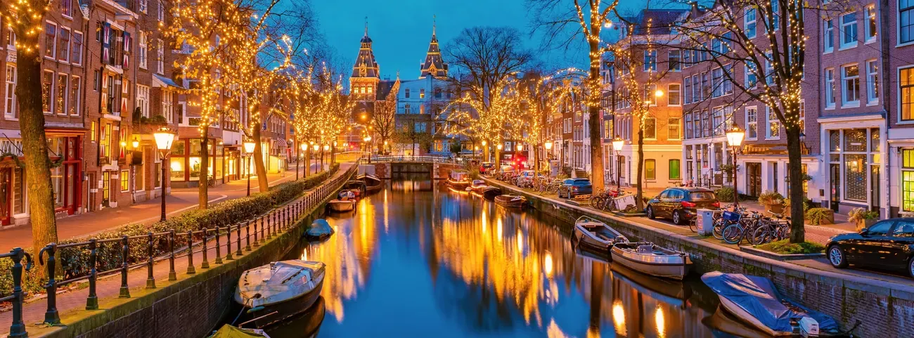 Christmas lights in the trees reflected in a canal in Amsterdam in the evening, with moored boats and the Rijksmuseum in the distance