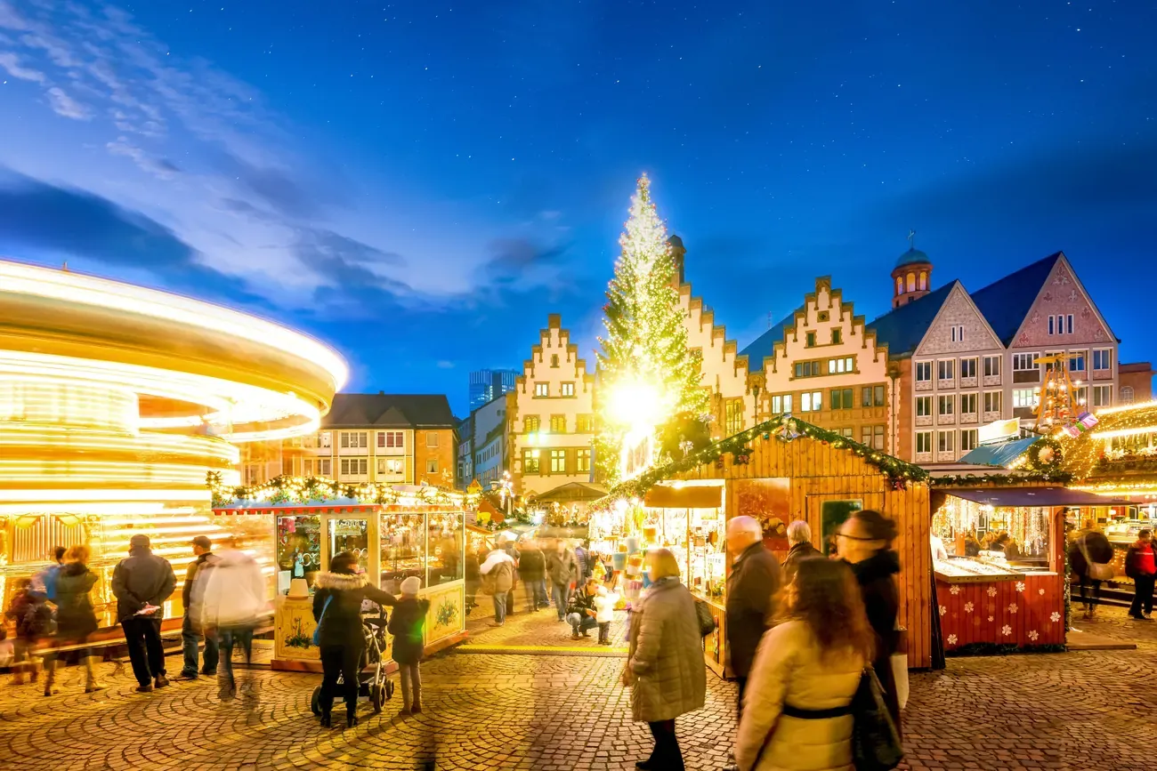 A spinning carousel beside lit stalls on Römerberg in Frankfurt, with the Christmas tree and the Römer gables behind