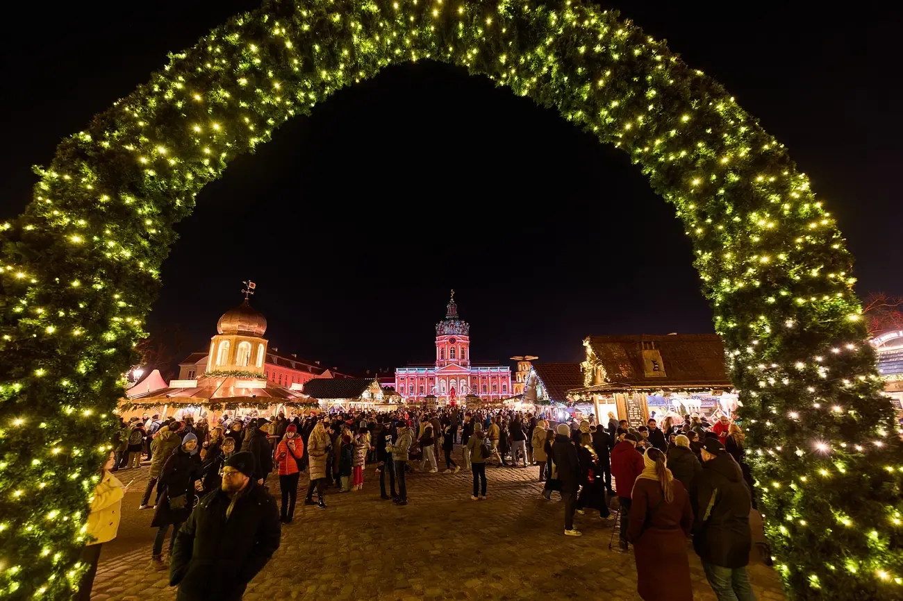 A lit arch of greenery framing the Christmas market and Charlottenburg Palace at night, Berlin
