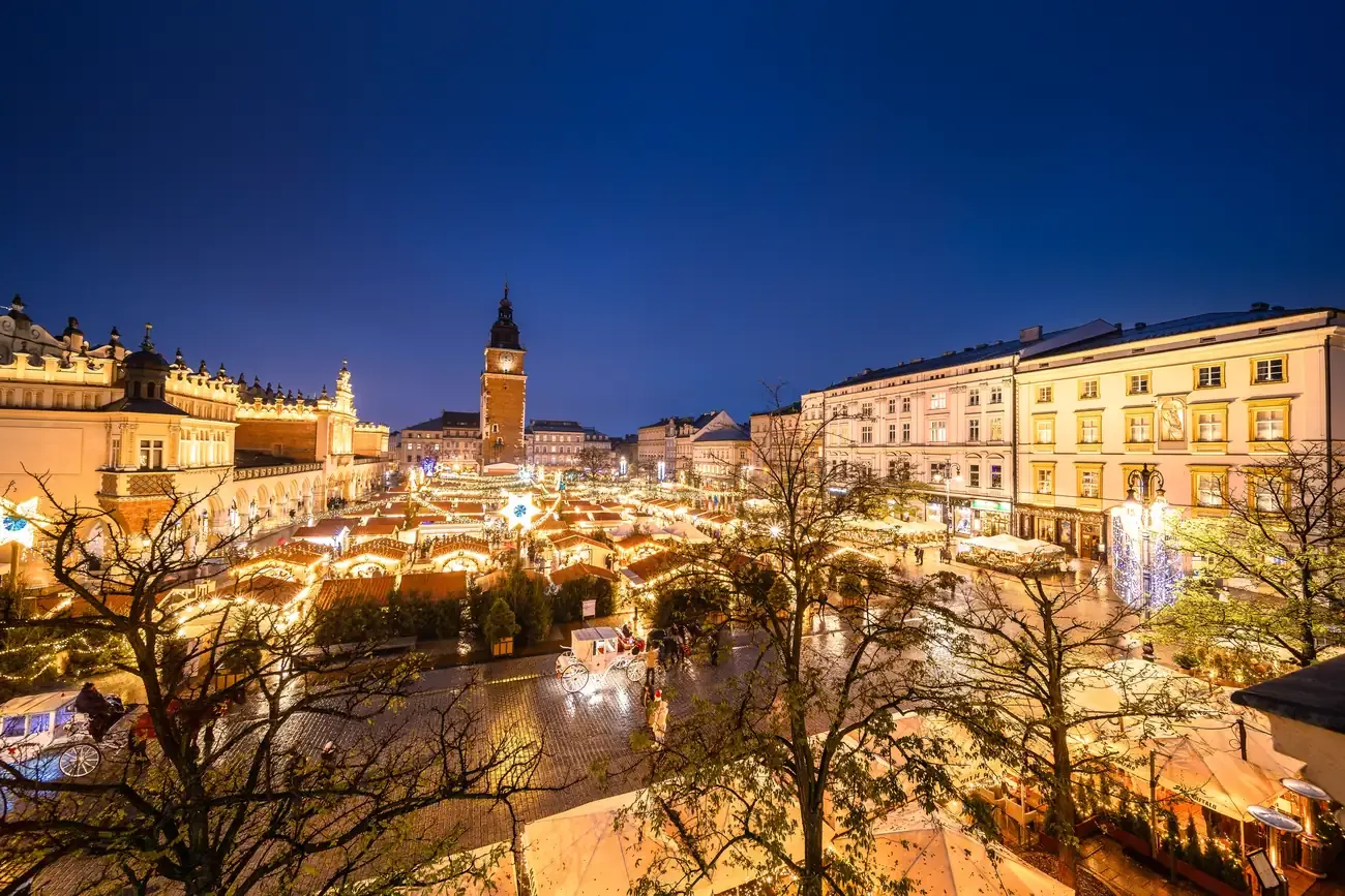 Lit huts and trees of the Christmas market on the Main Market Square in Krakow, with the Town Hall Tower and the Cloth Hall behind