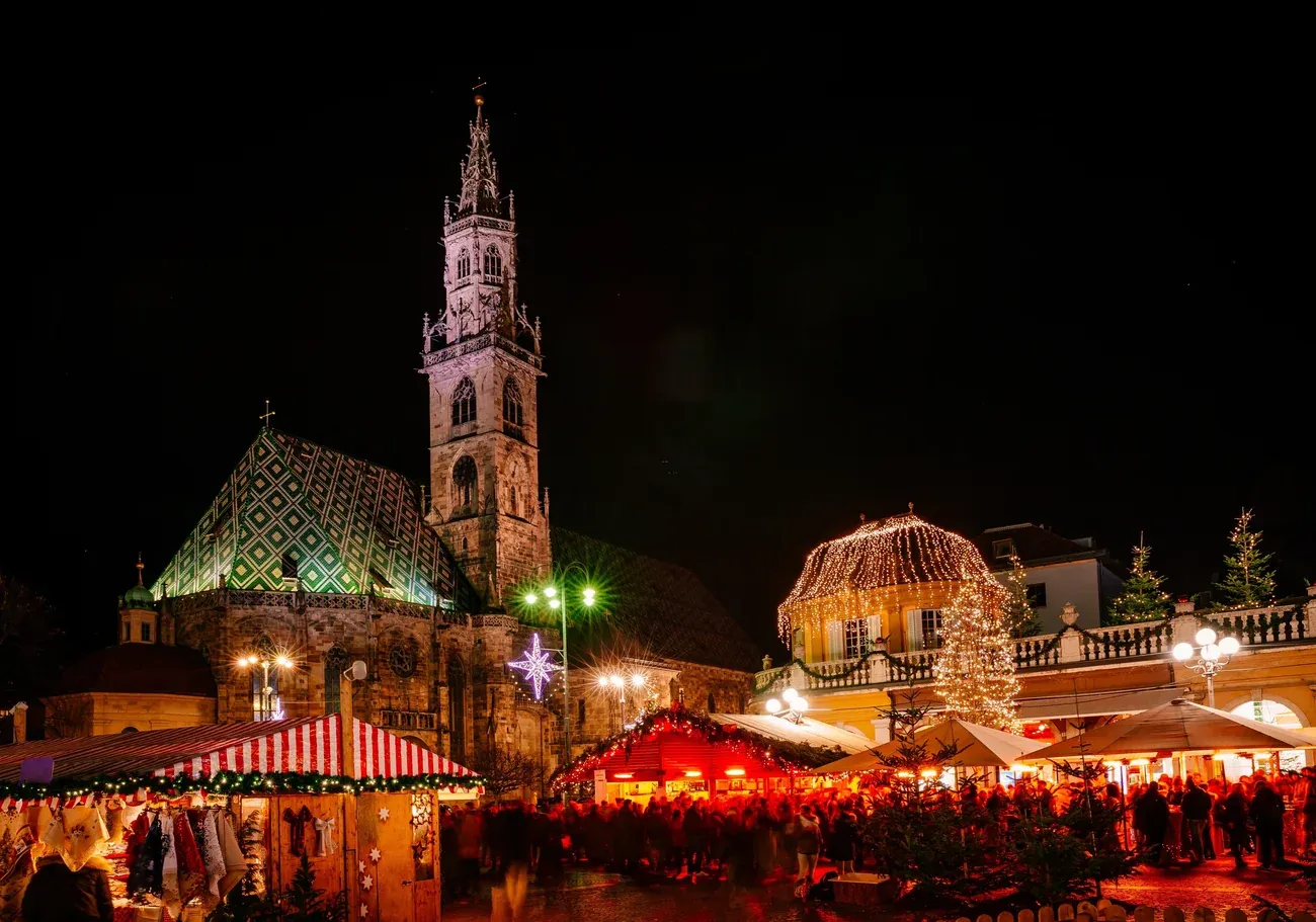 Christmas market stalls on Piazza Walther in Bolzano at night, with the cathedral's patterned roof and spire behind