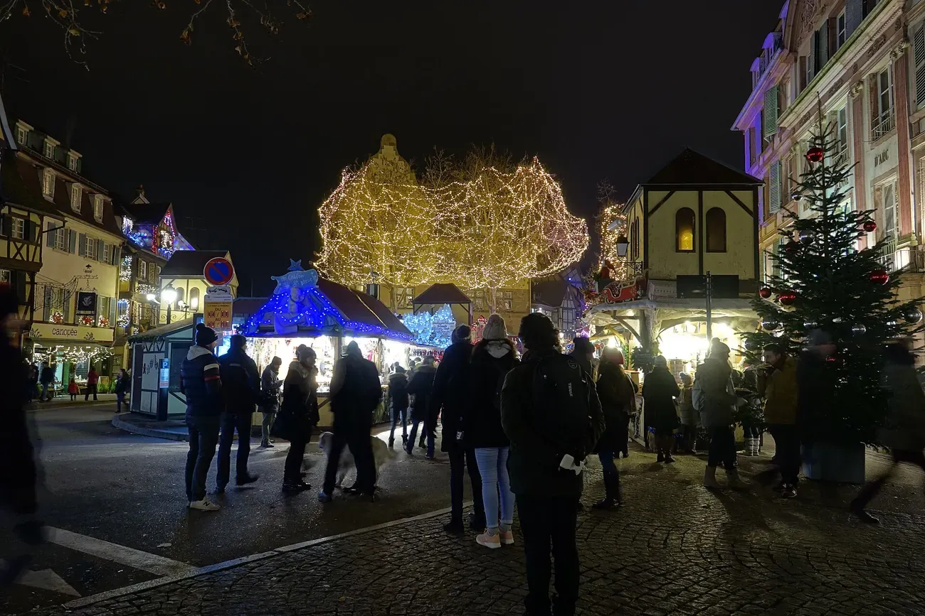 Visitors among the chalets and lit trees of the Christmas market on place Jeanne d'Arc in Colmar at night