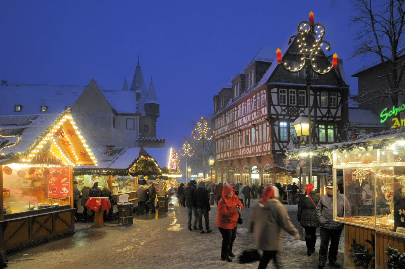 Lit stalls and visitors on Römerberg in Frankfurt at the Christmas market, with snow on the roofs and half-timbered houses behind