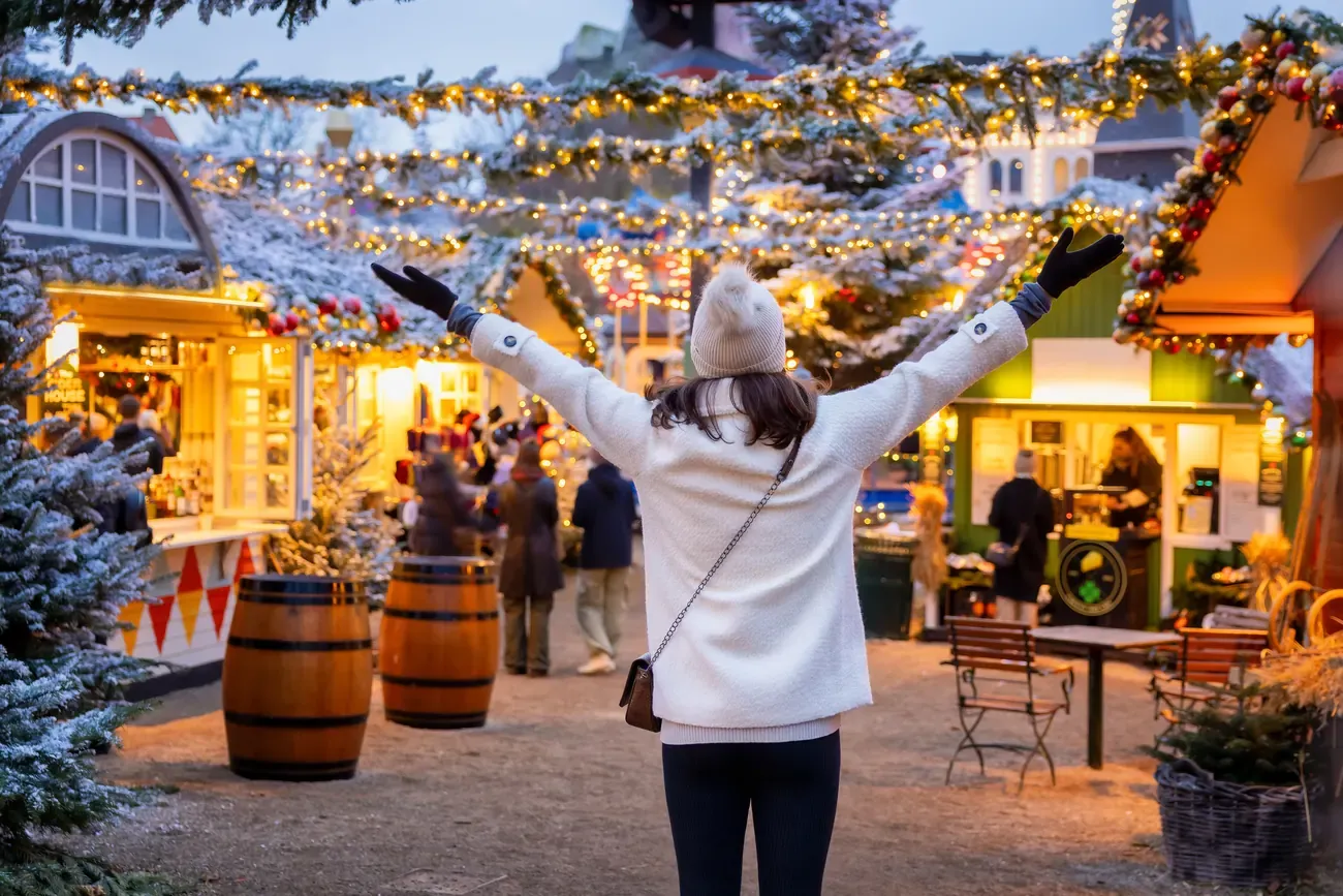 A visitor with outstretched arms among lit Christmas stalls and snowy fir trees in Tivoli Gardens, Copenhagen, at dusk