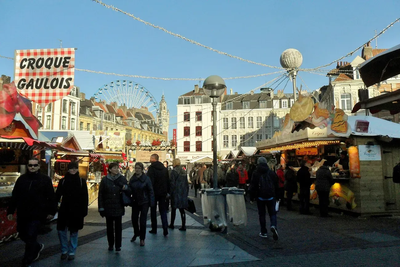 Visitors between Christmas market stalls in Lille, with a big wheel and the belfry of the Chamber of Commerce behind