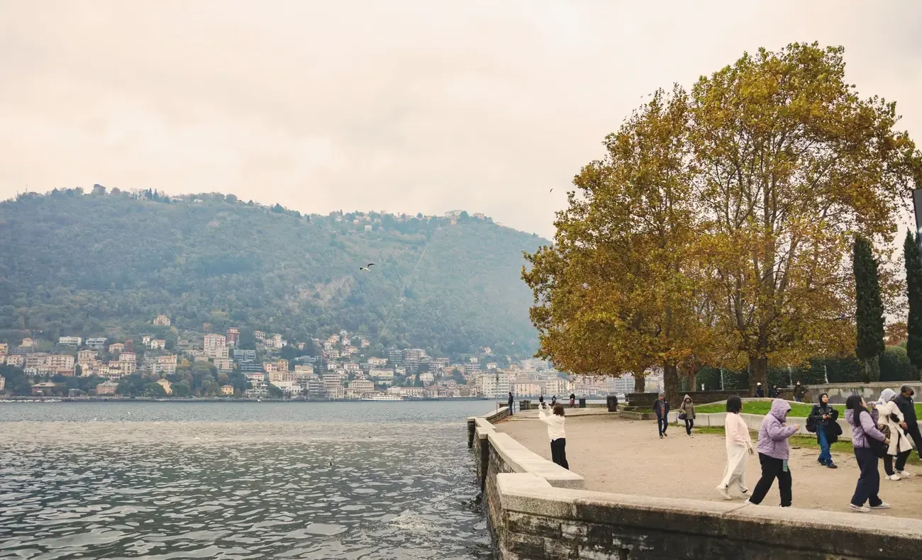 Walkers on the stone lakefront promenade in Como in autumn, with Brunate hill across the water