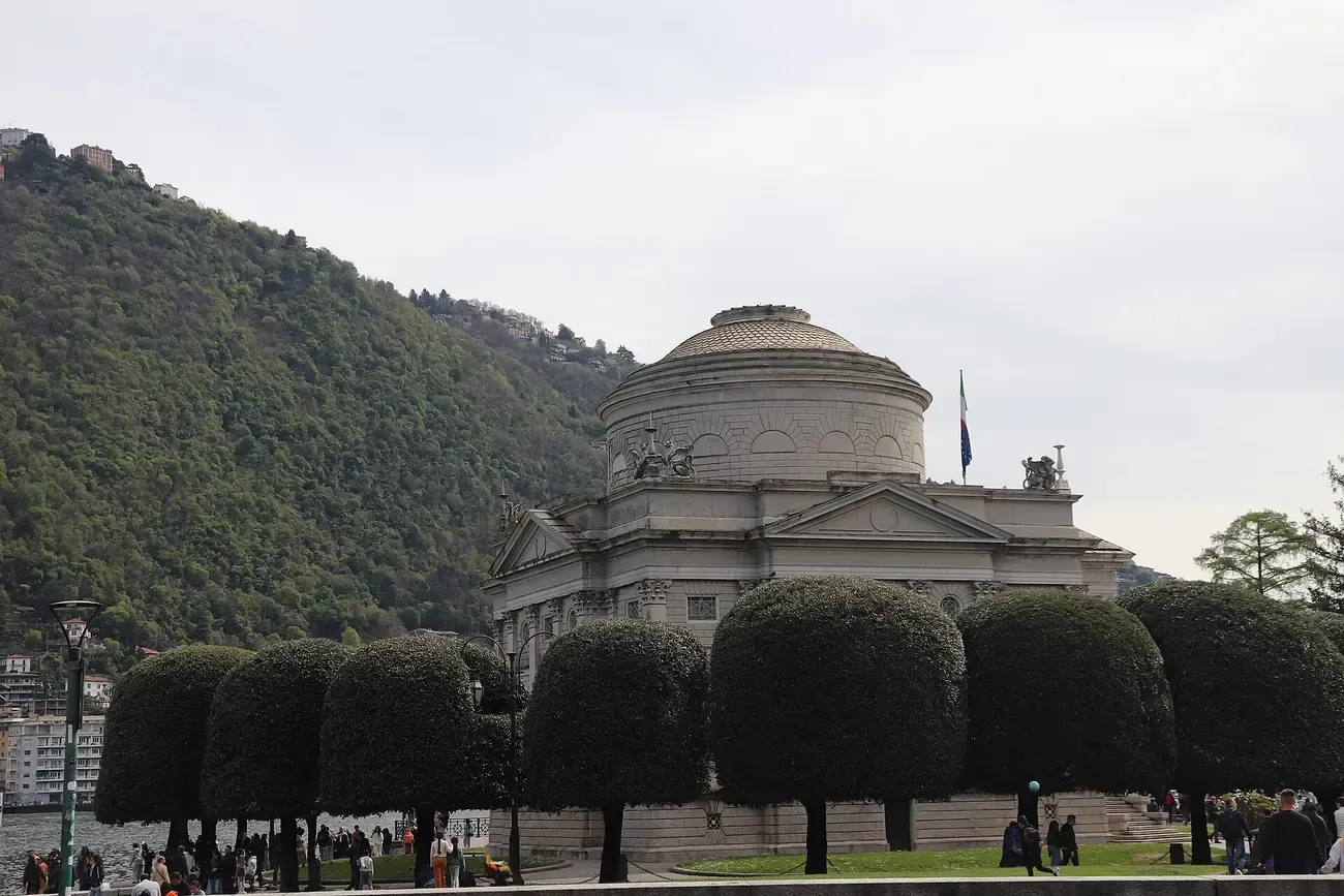 Side view of the domed Tempio Voltiano in Como behind clipped trees, with the hill behind
