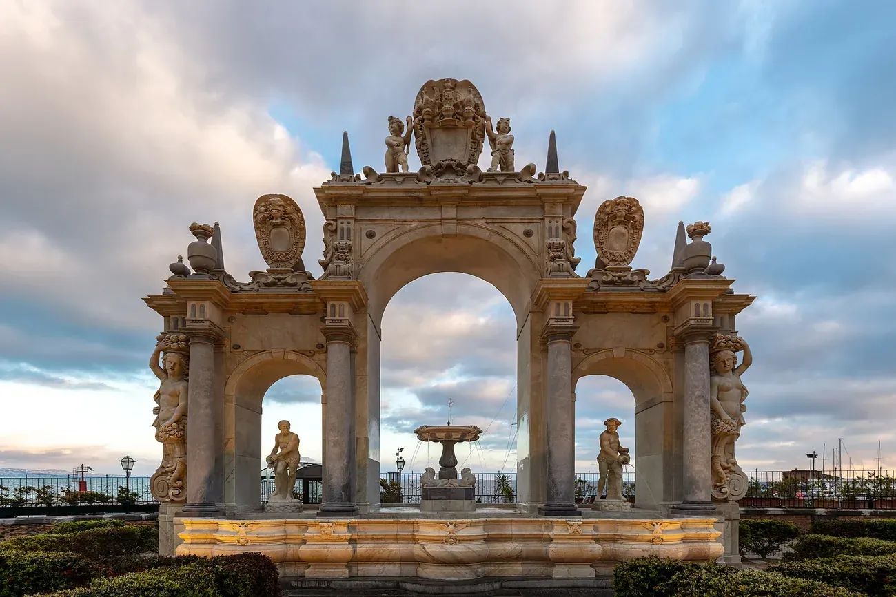 The three-arched marble Fontana del Gigante on the seafront of Naples at dusk with the bay behind