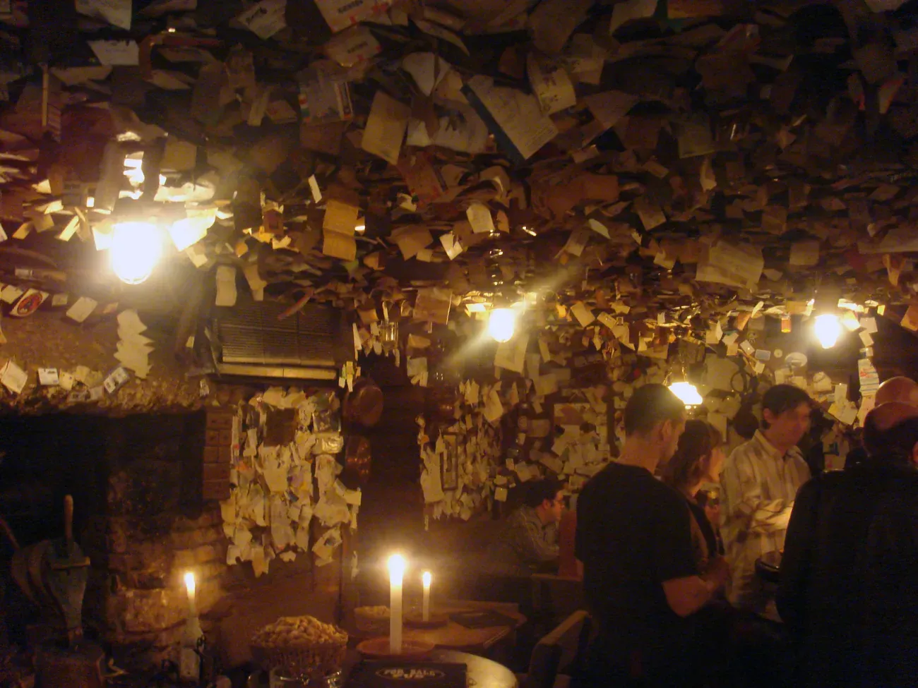The candlelit interior of the For Sale Pub in Budapest, with walls and ceiling covered in notes and business cards