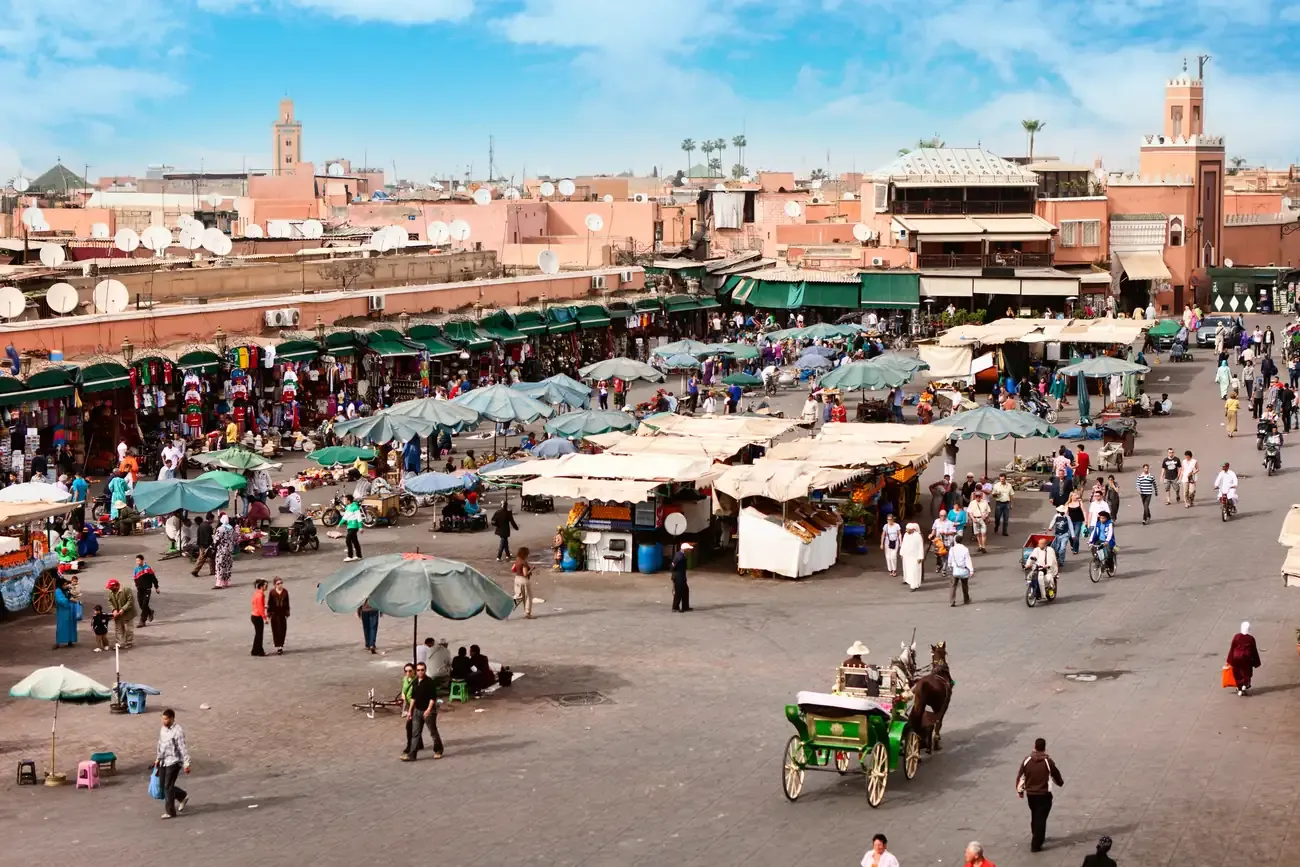 Daytime aerial view of Jemaa el-Fnaa square filled with white market stalls and a horse carriage, with the Koutoubia minaret in the distance, Marrakech.