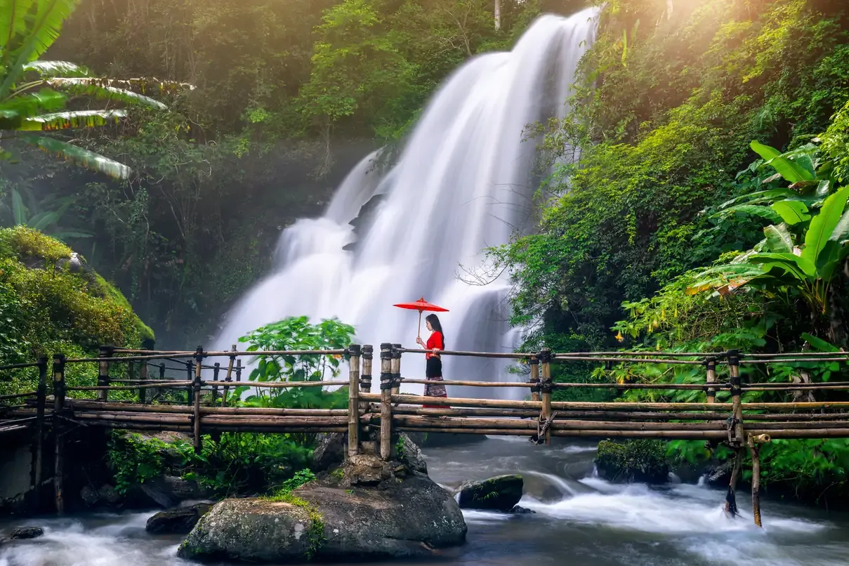 A woman with a red umbrella on a bamboo bridge before a jungle waterfall, Chiang Mai