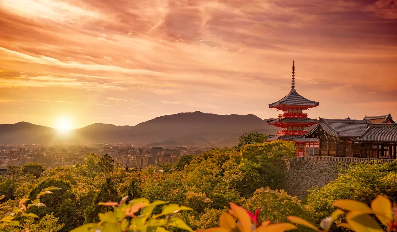 The three-storey pagoda of Kiyomizu-dera above the treetops with the sun setting over Kyoto