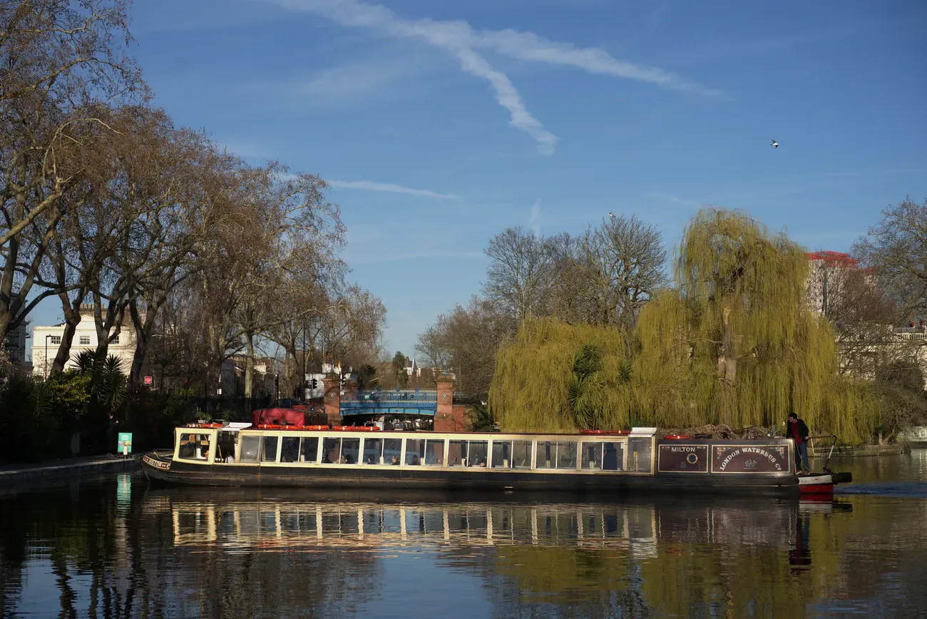 Narrowboat on the water at Browning's Pool in Little Venice, with willows on the island and moored boats beyond, London