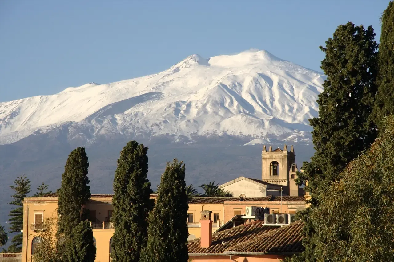 The snow-capped summit of Mount Etna rising above the rooftops and cypresses of Taormina in Sicily