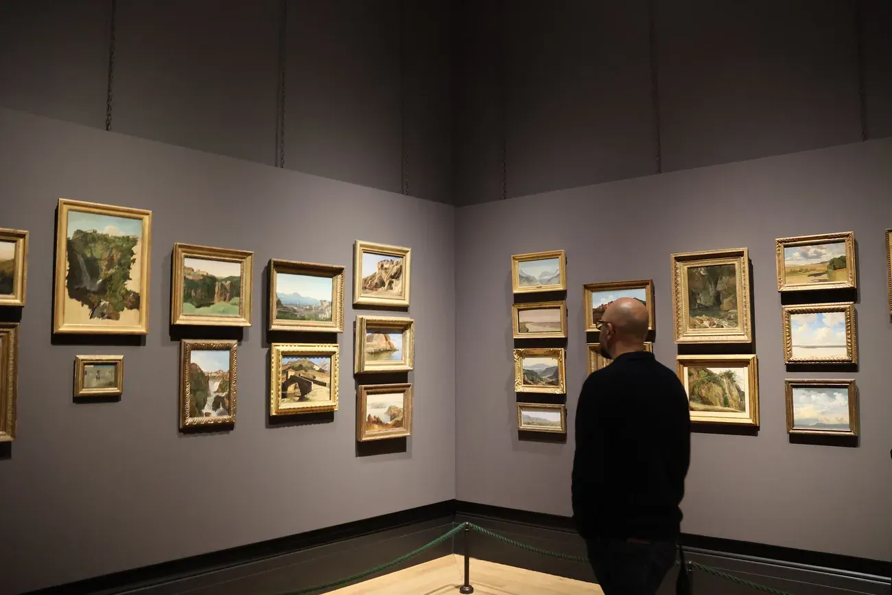 A visitor looking at landscape paintings in a grey-walled room of the National Gallery, London