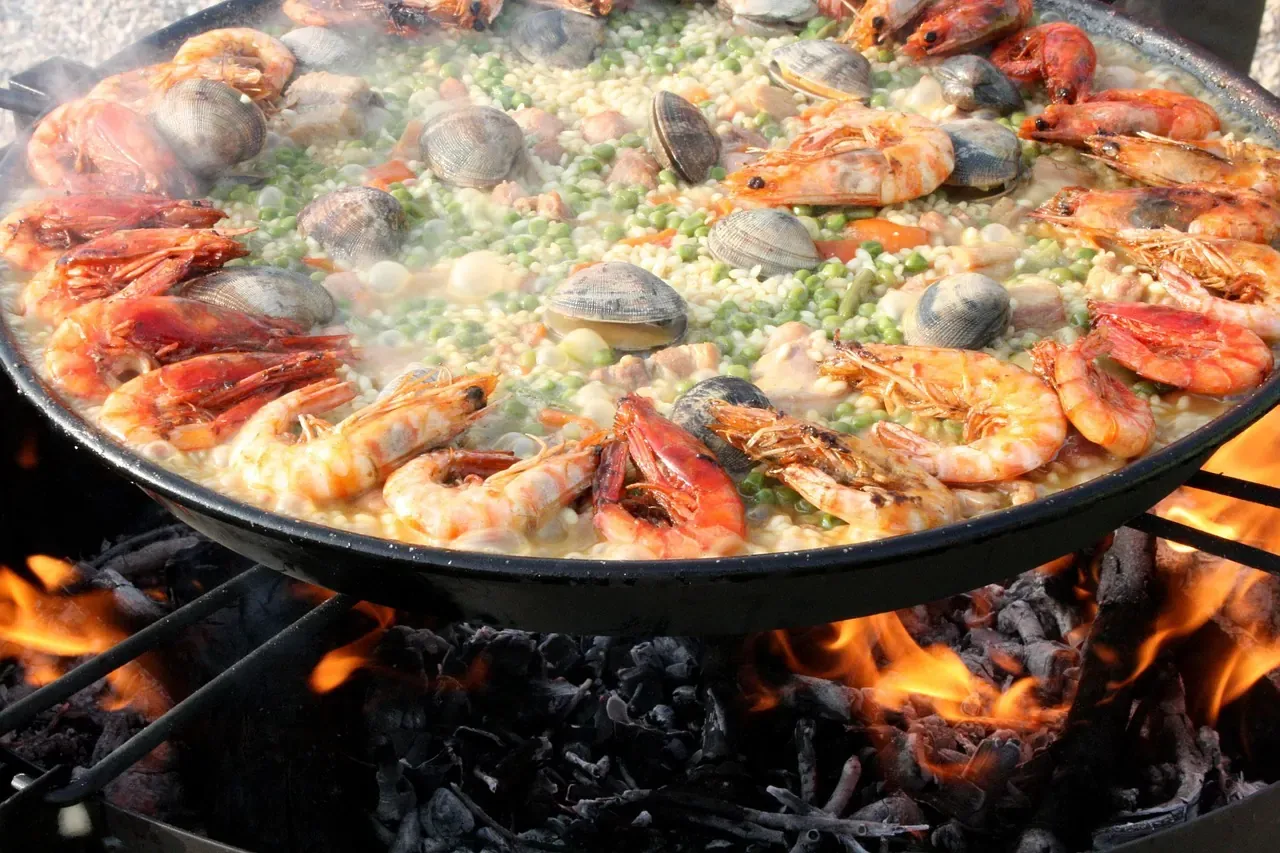 A wide paella pan of rice with prawns and clams cooking over an open fire