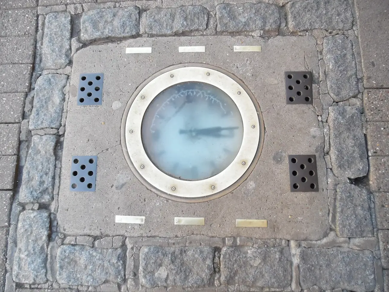 A round clock set into the paving of Thames Street in Windsor, framed by brass strips and ventilation plates