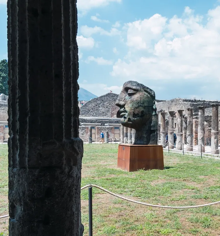 Monumental bronze head sculpture standing among the colonnades of the Pompeii ruins