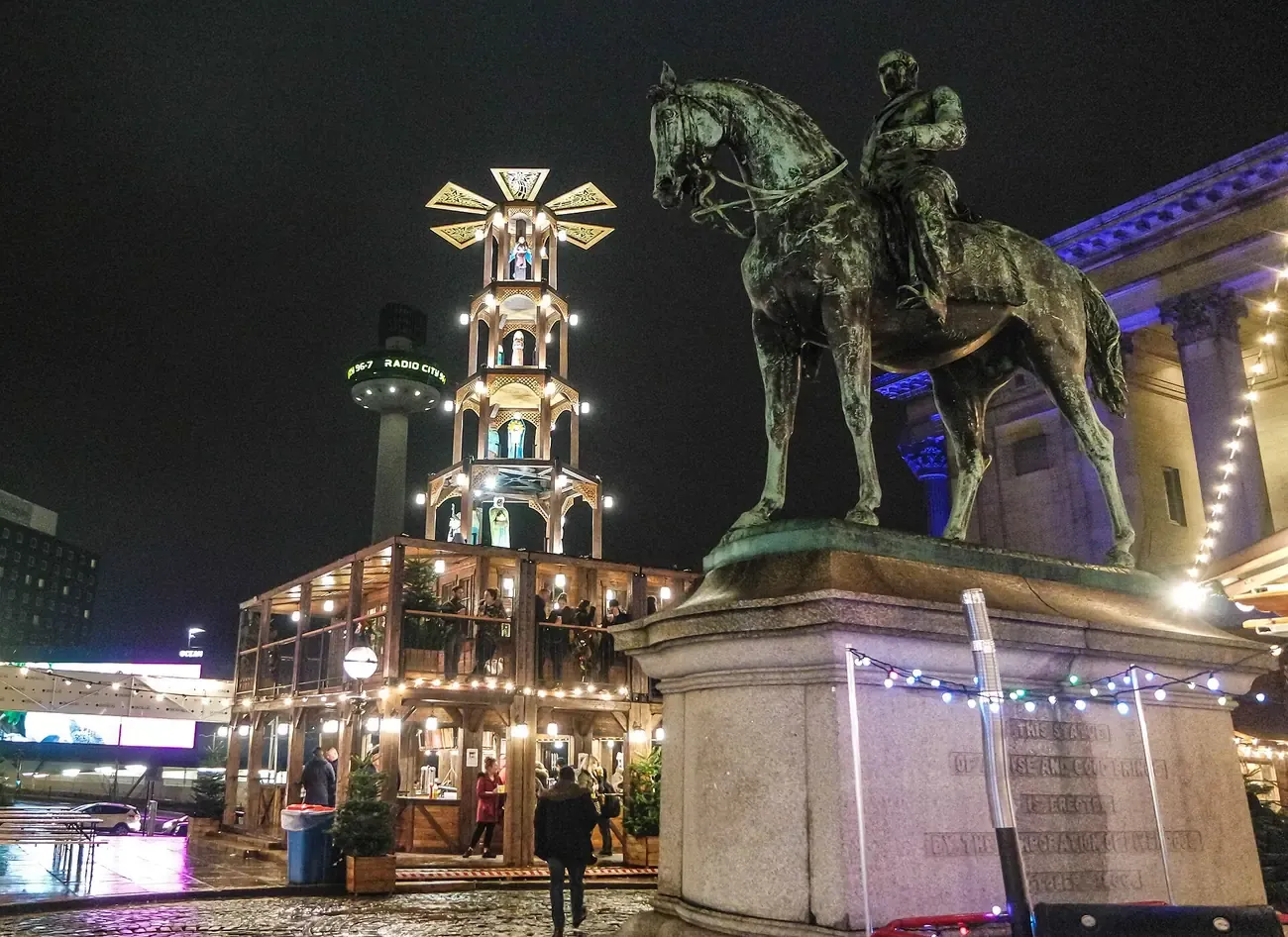 The equestrian statue of Prince Albert on St George's Plateau in Liverpool at night, with lit Christmas chalets and a tiered wooden tower behind