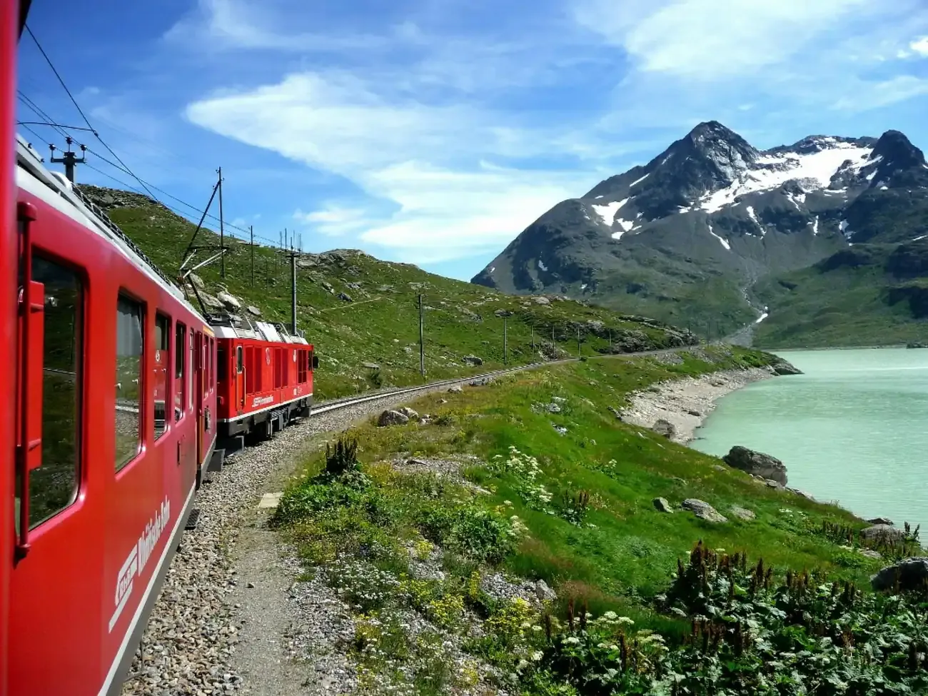 Red Bernina train running beside an Alpine lake with a snow-capped peak under a blue sky