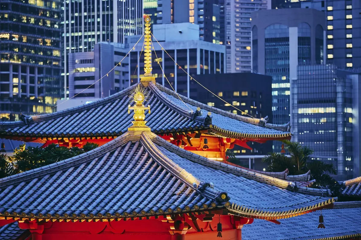 Golden finial and blue tiled roof of a Buddhist temple in Singapore standing against lit office towers at twilight