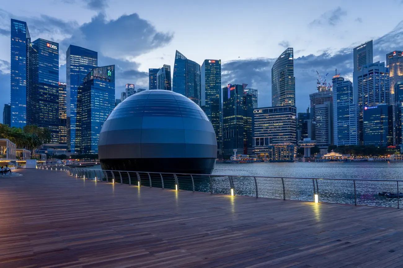 Wooden boardwalk promenade beside the glass dome pavilion on Marina Bay with the financial district skyline behind at blue hour in Singapore