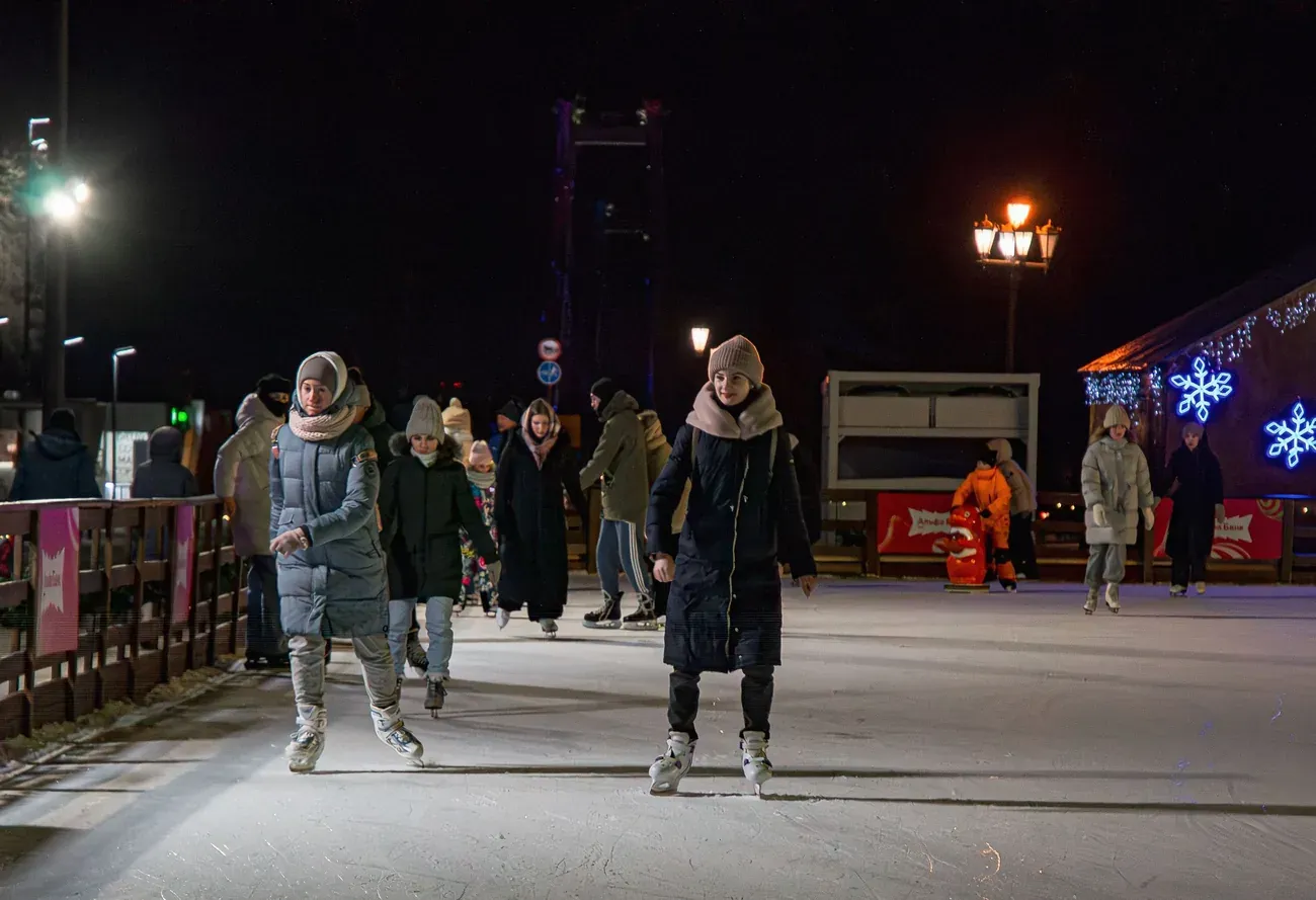 Skaters on a floodlit outdoor ice rink with festive lights at night