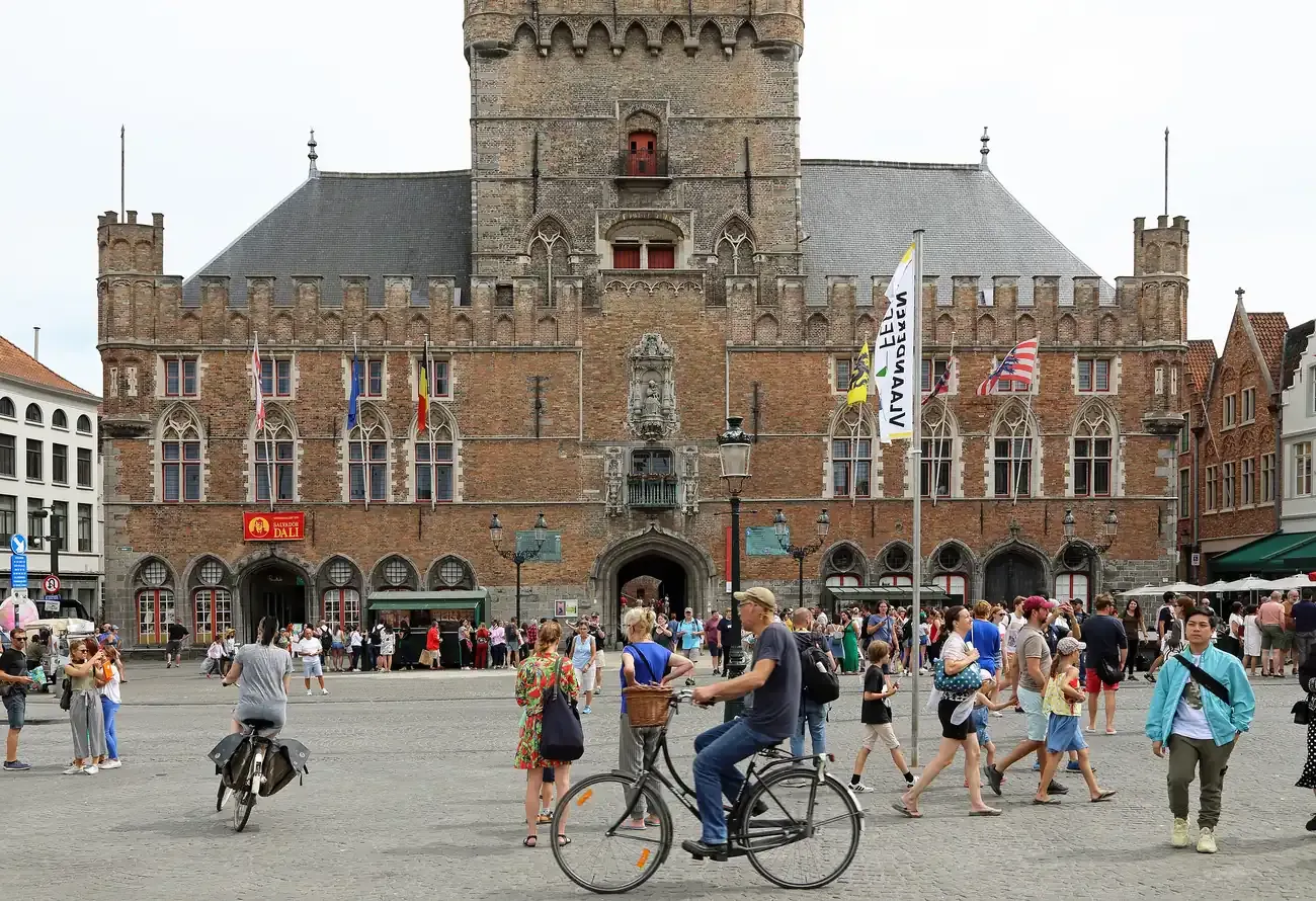 The brick front of the Stadshallen at the foot of the Belfry on the Markt in Bruges, with people and cyclists crossing the square