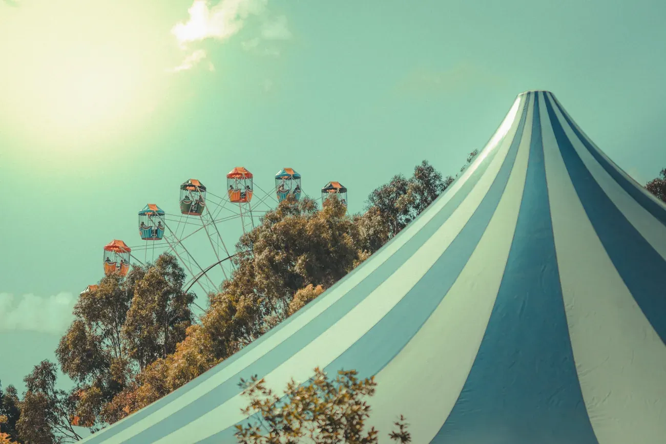 Blue and white striped circus big top beside a Ferris wheel and trees