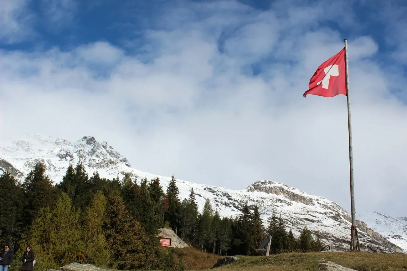 Swiss flag flying above pine forest and snow-covered Alpine peaks