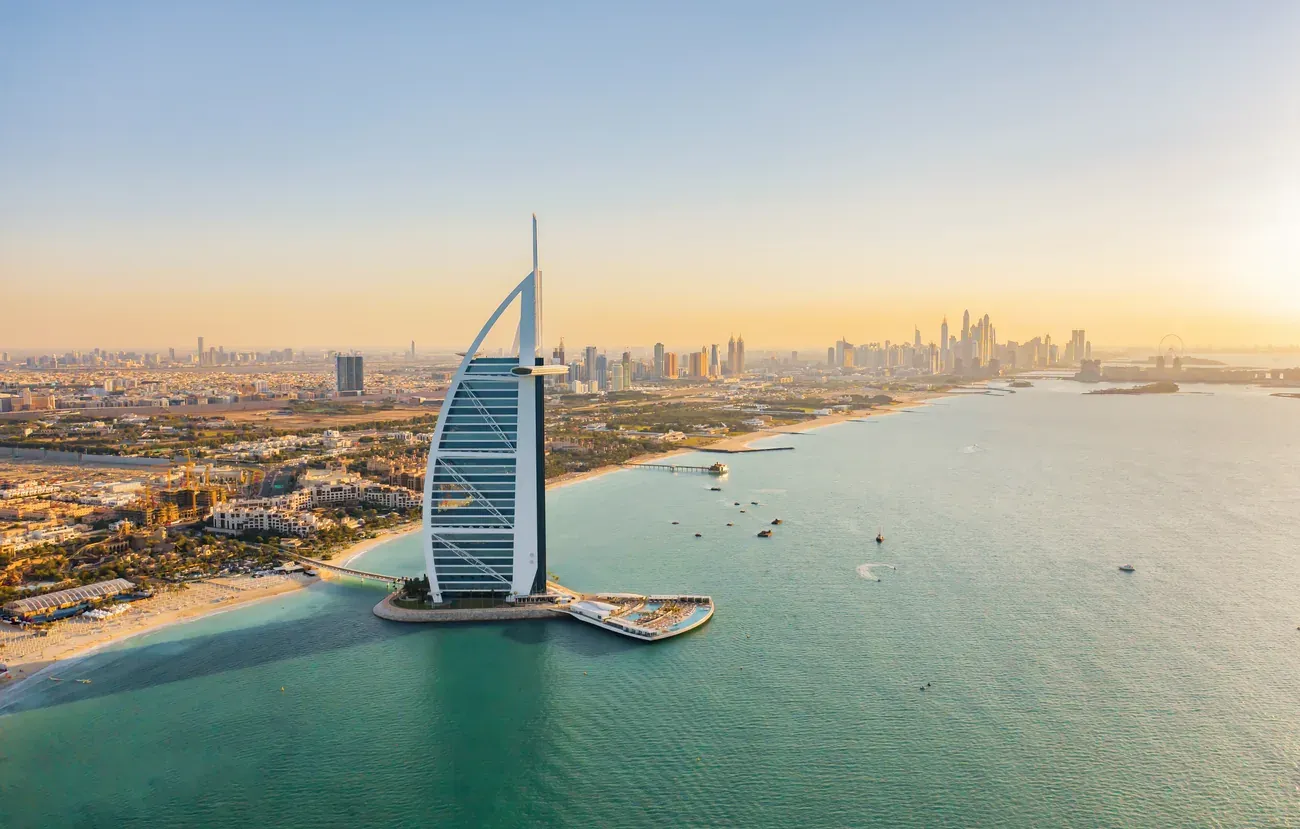 Aerial view of the Burj Al Arab with the Dubai Marina skyline in the distance.