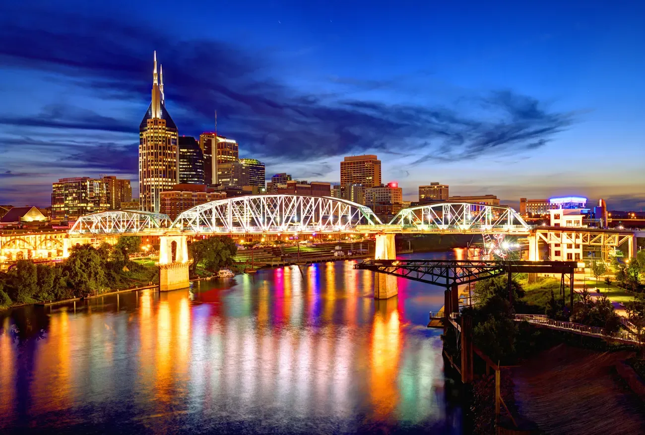 Nashville skyline reflected in the river beside the lit pedestrian bridge at night