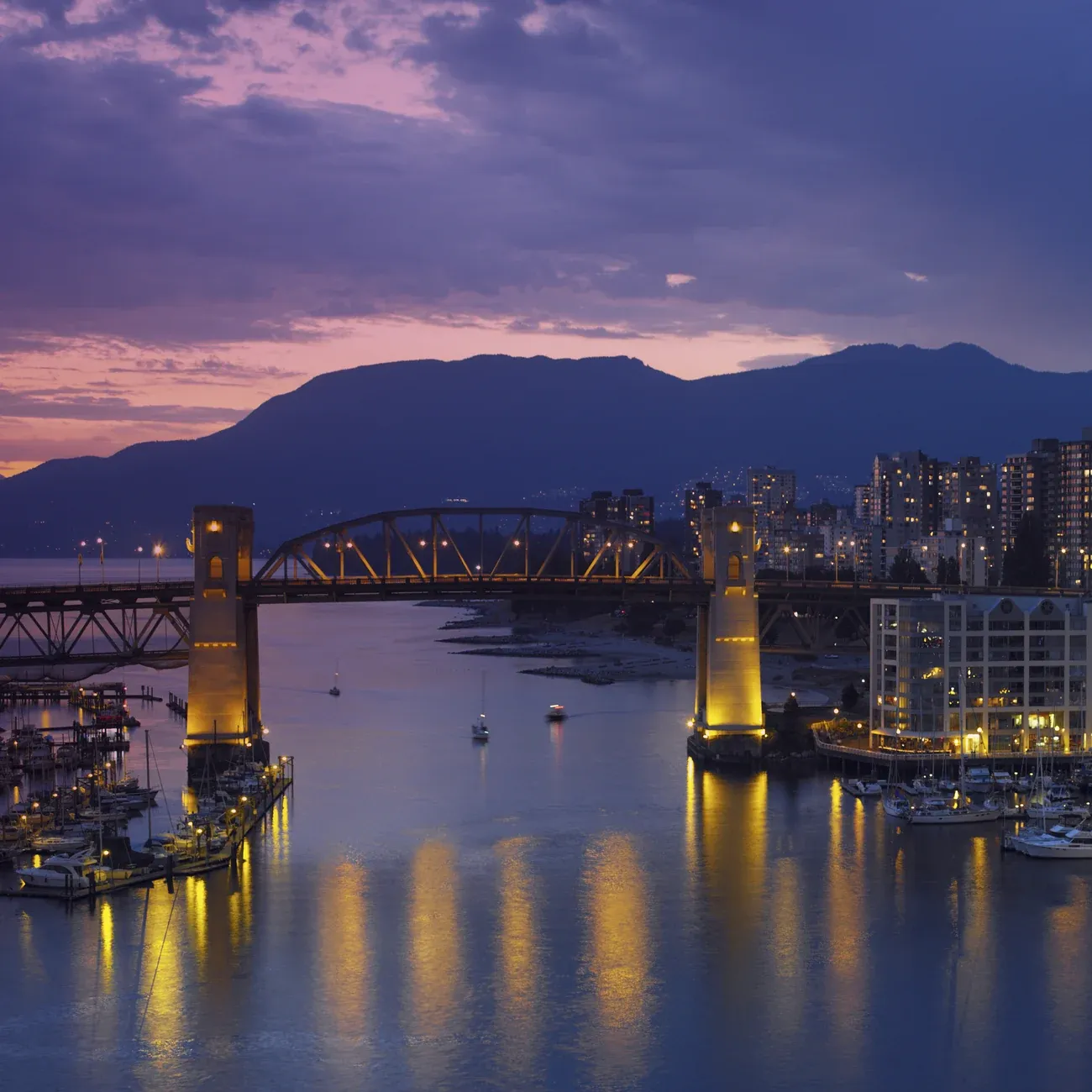 Burrard Street Bridge lit above False Creek and its marinas in Vancouver at dusk, with the North Shore mountains behind