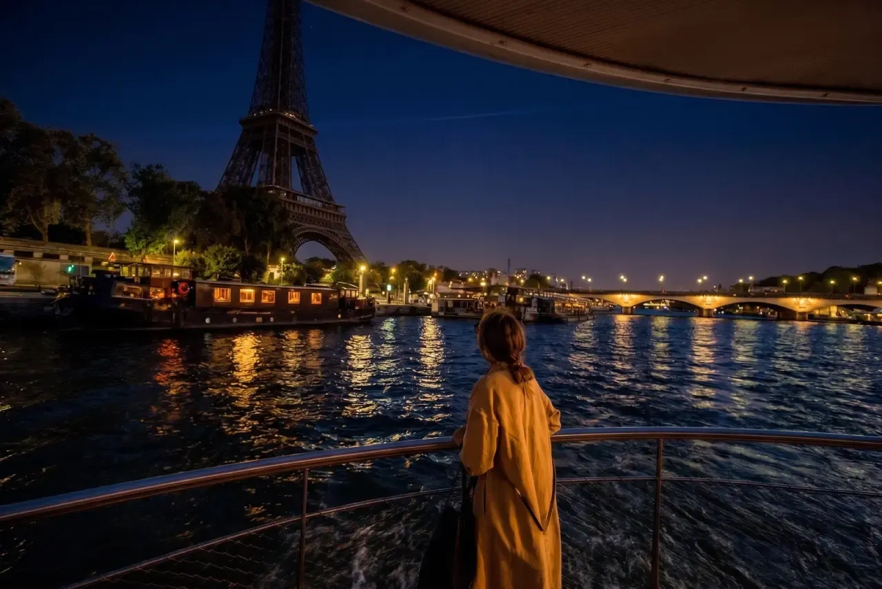 A woman watching the Eiffel Tower at night from a Seine river cruise