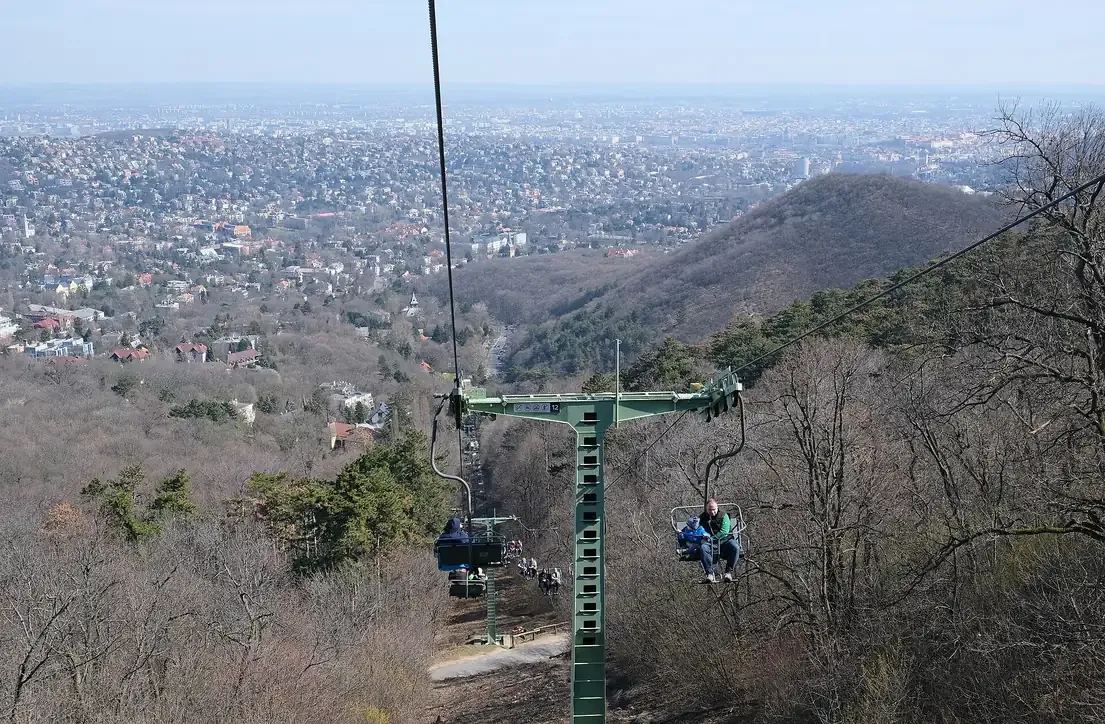 Open chairs of the Zugligeti chairlift climbing through the bare treetops of the Buda hills with the Budapest skyline behind